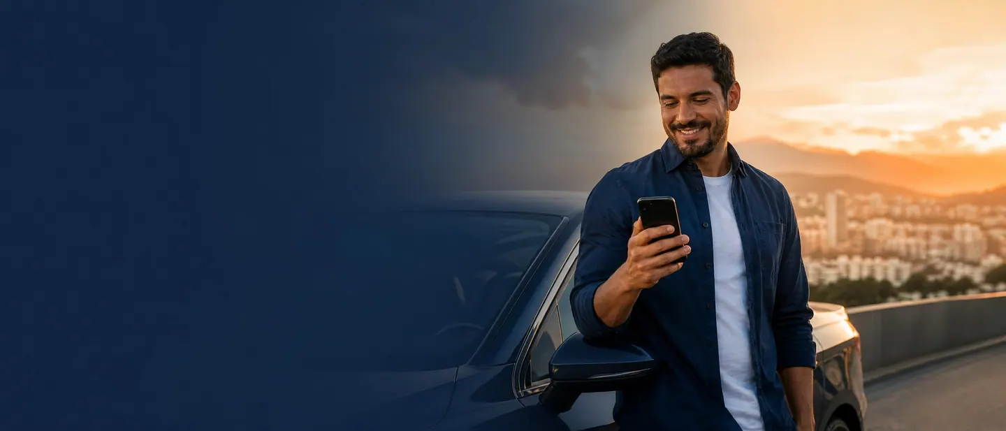 Homem sorrindo ao conferir o celular, encostado no próprio carro em um mirante ao pôr do sol.