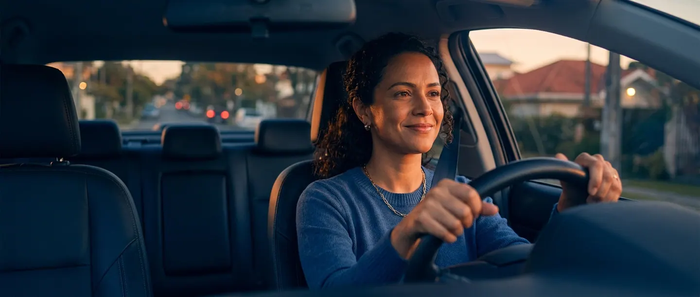 Mulher ao volante do próprio carro, sorrindo em uma rua tranquila no fim da tarde.